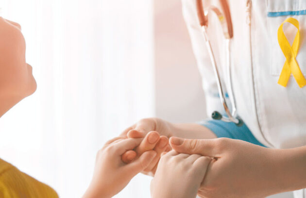 Criança olhando para um médico enquanto segura sua mão, o qual tem uma fita de luta contra o câncer em seu jaleco